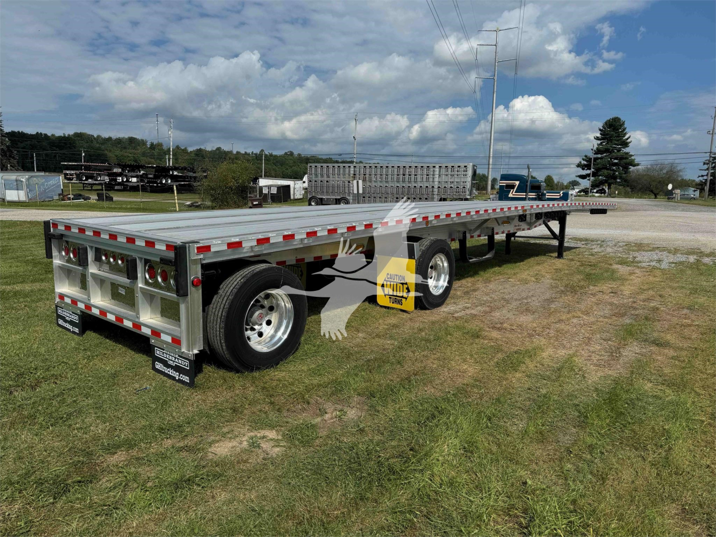 Flatbed trailer showing dual rear axles and aluminum decking with reflective tape; rear lights and mud flaps visible; stock tag and branding on tires.