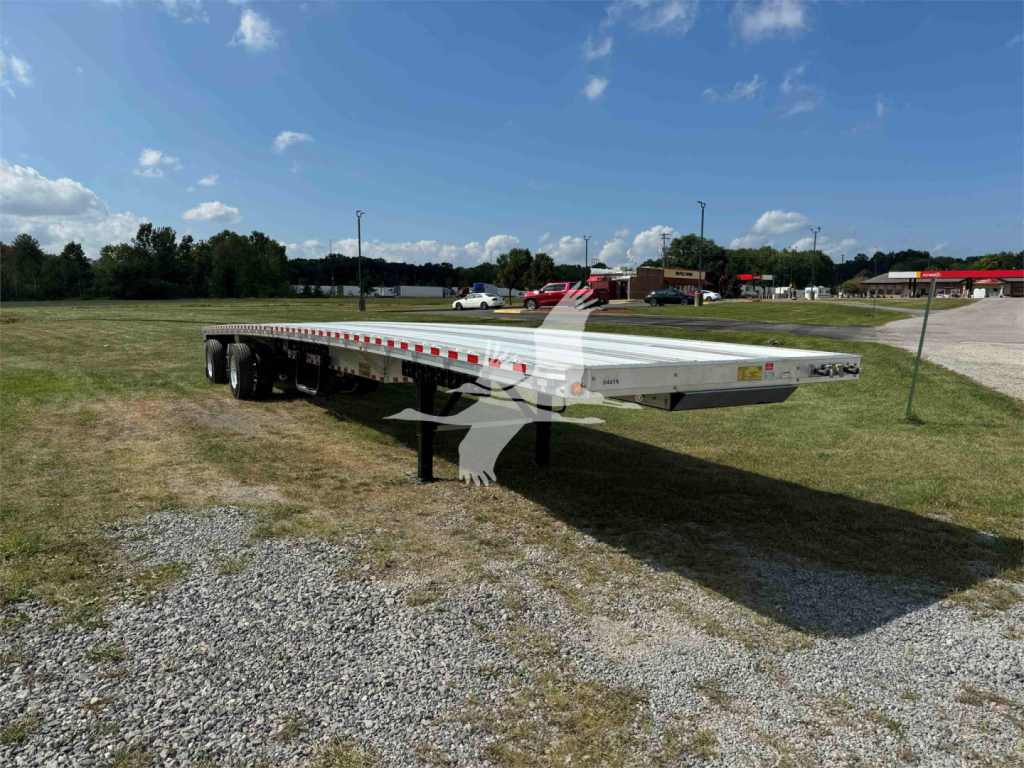 Trailer bed showing a long flatbed trailer with dual rear axles and kickstand, silver finish, no cargo, outdoor lot.