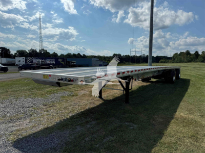 Flatbed trailer in side view showing dual axles and extended deck with reflective tape and support legs; sky and grassy field background.