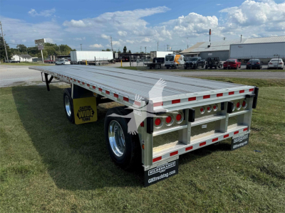 Flatbed trailer showing rear with dual axles and silver toolbox, cargo deck with metal planks, labeled reflectors, and signage in back; 2025 Reitnouer Maxmiser trailer.