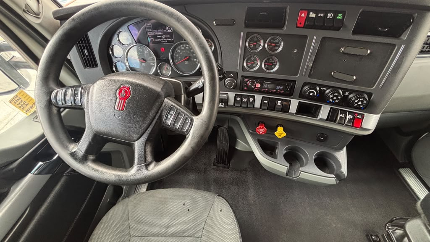 Kenworth T680 cab interior showing steering wheel, gauge cluster, and dashboard controls with pedals and center console.