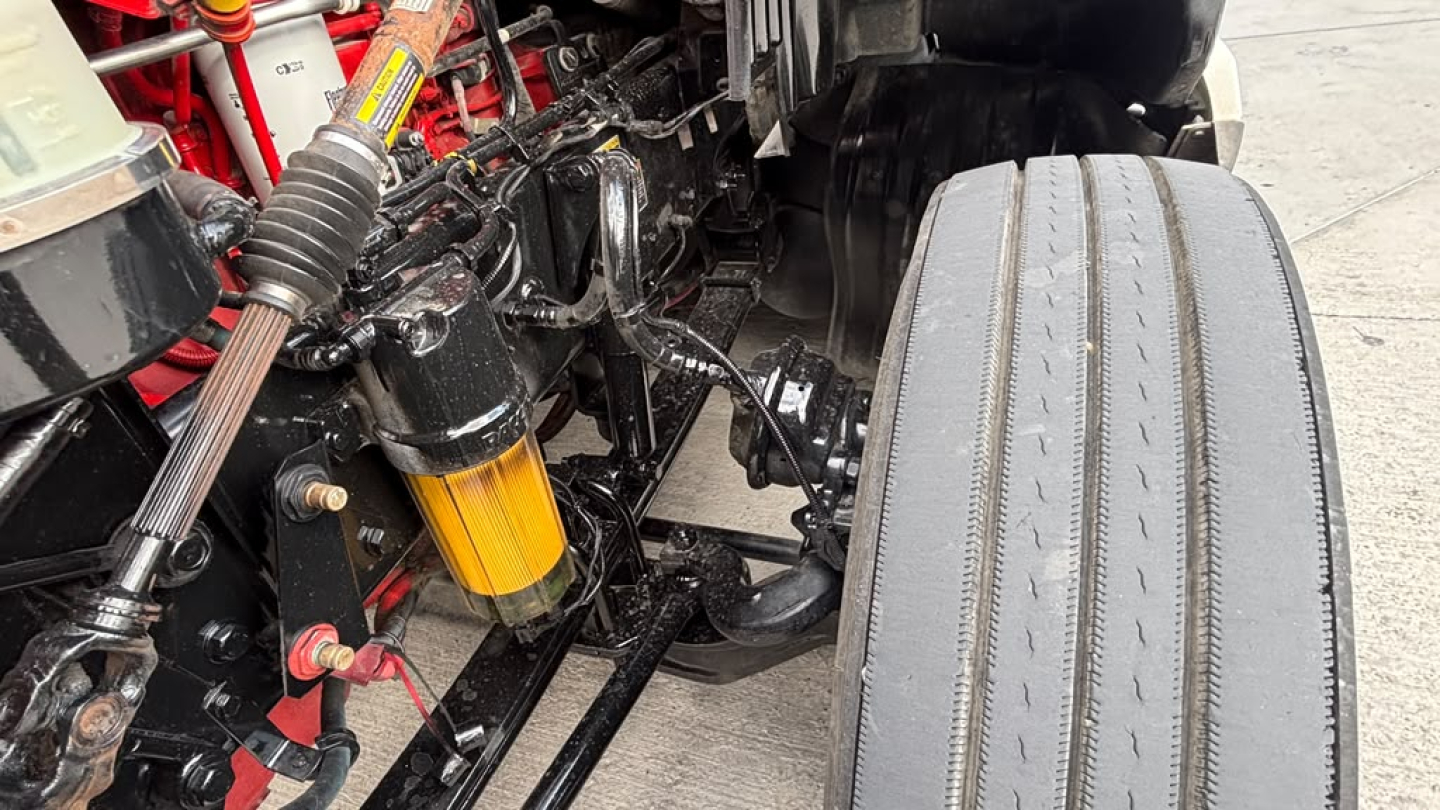 Kenworth T680 chassis undercarriage showing suspension components, air lines, and a single wide tire. Visible are yellow shock absorber, red-painted frame, and various hoses with metal fittings.