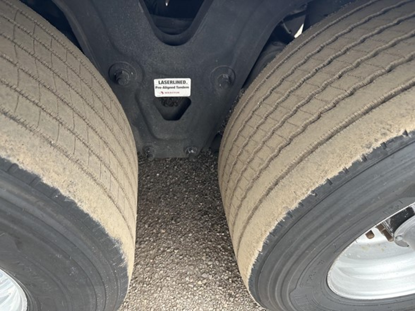 Trailer undercarriage showing dual tires and suspension with a small label reading 'LASER LINED' on the chassis. Visible part of the fifth wheel area and ground surface between wheels.