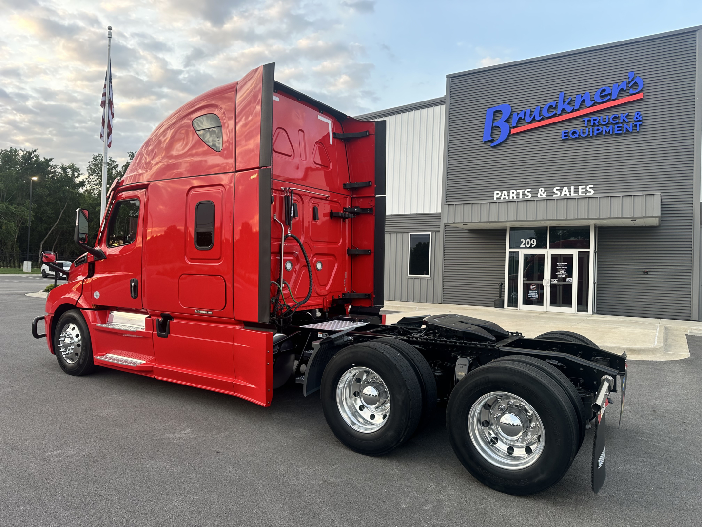 Truck cab of a red Freightliner Cascadia 126 tractor seen from the side with sleeper cab and dual rear axles, showroom building in background.