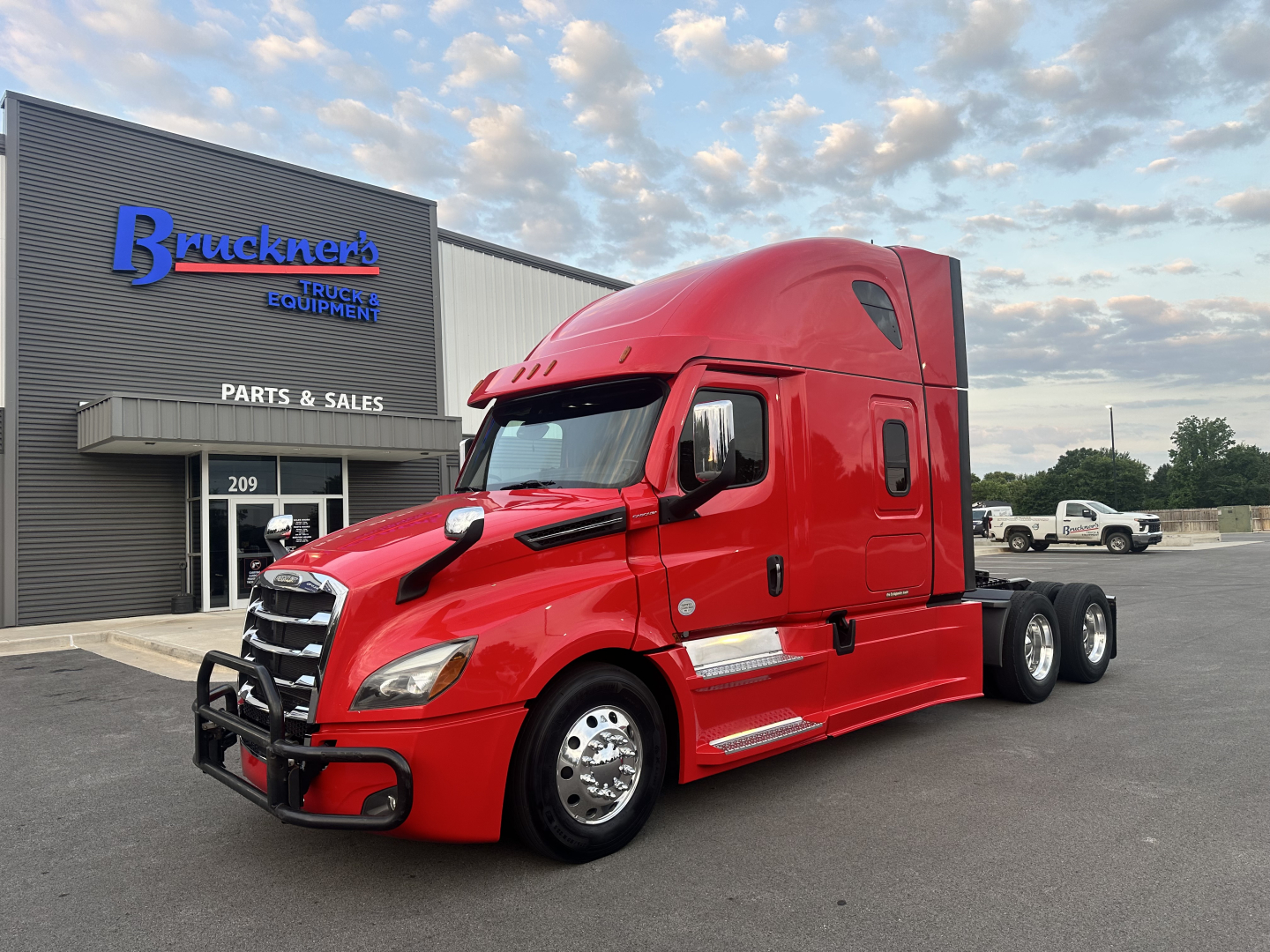 Freightliner Cascadia 126 tractor in red parked outside a service building with blue 'Bruckner's' sign; text on building reads PARTS & SALES and 209 door; no other labels clearly visible.
