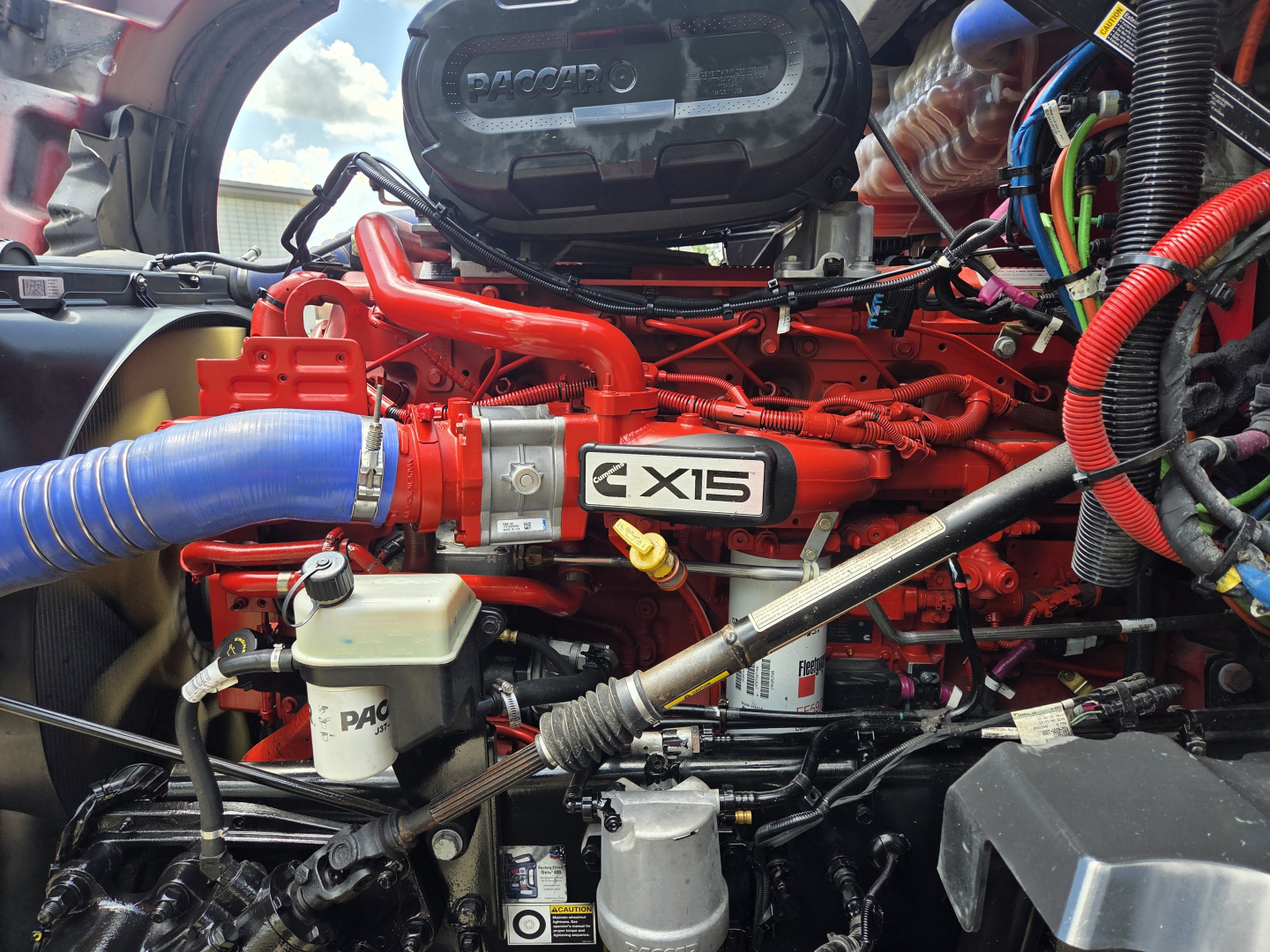 Kenworth T680 engine bay with red engine and X15 badge, blue radiator hose, various lines and wiring visible.