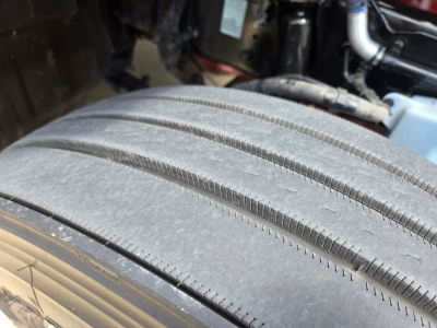 Kenworth T680 wheel and tire close-up showing tread and sidewall with engine bay components faint in background.