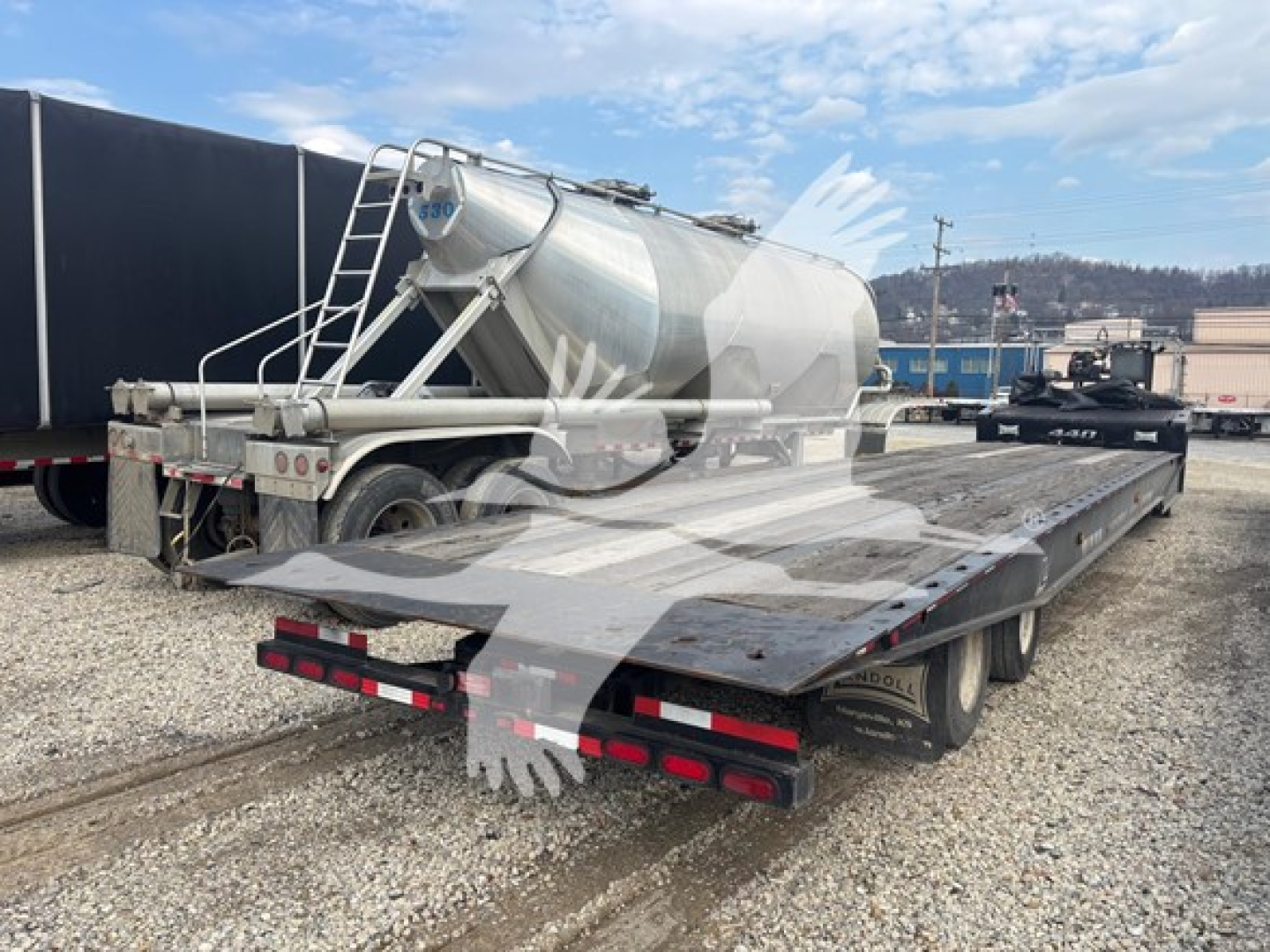 Flatbed trailer with a cylindrical stainless steel liquid transport tank mounted on a dual-axle bed; rear license plate area visible; side ladder and railing on tank.