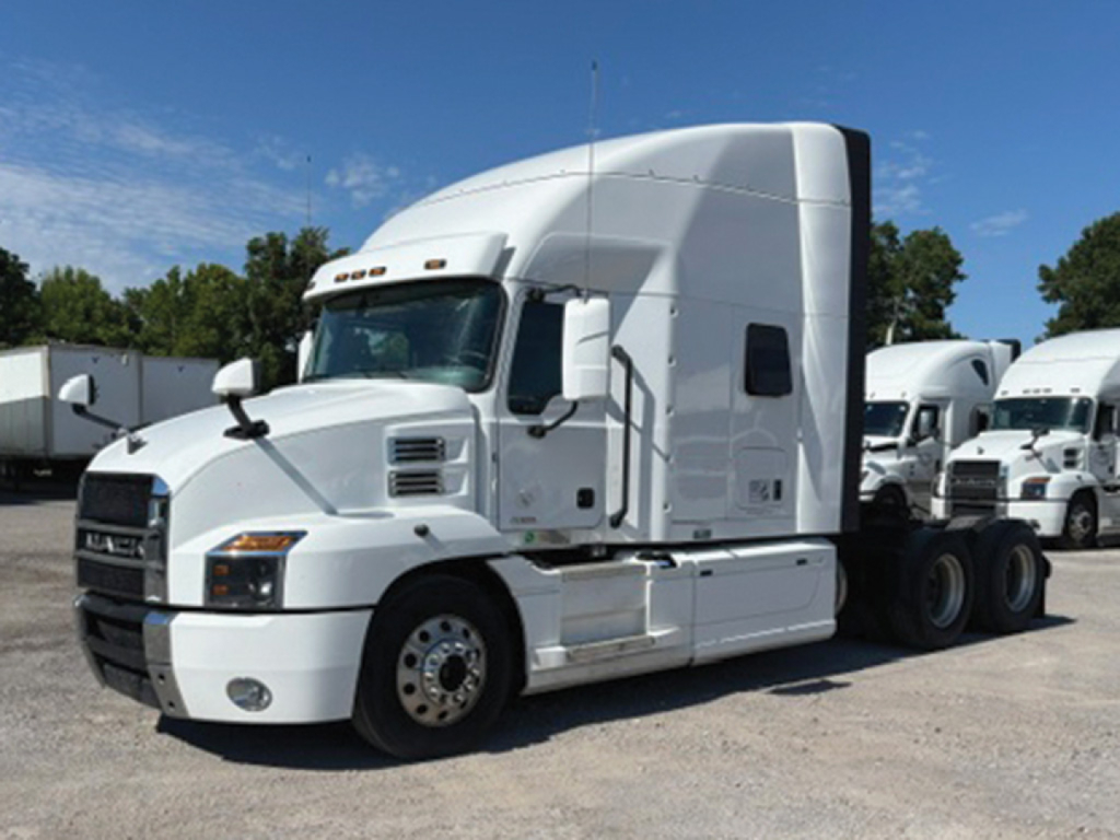 Truck cab with sleeper on a Mack Anthem showing white exterior and front grille, side mirrors, and attached trailers in background.