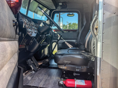 Peterbilt 379 truck cabin interior showing driver seat and dashboard with steering wheel, gear shifter, and control cluster; red fire extinguisher mounted under seat.