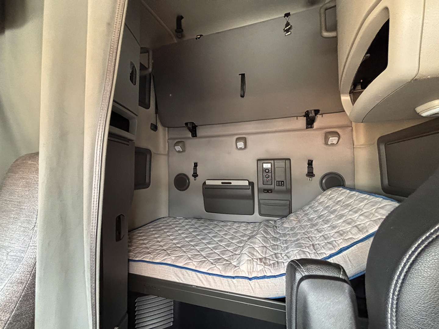 Truck sleeper cab interior with a single wide bunk mattress, control panel, storage compartments, and wall panels. No visible text legible.