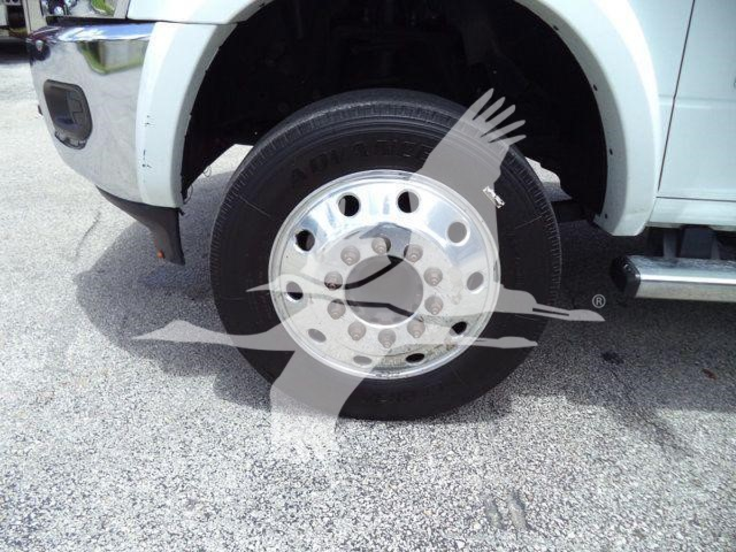 Wheel and front fender of a white Ram 5500 chassis cab showing a chrome multi-hole hubcap and tire, partial bumper and step rail visible.