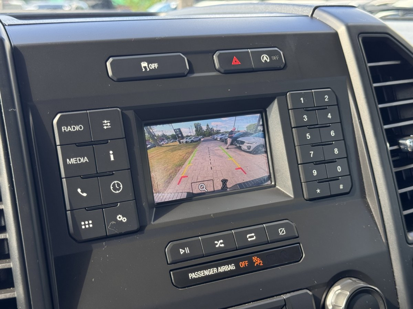 Ford F-150 center console with backup camera display showing parking grid and controls labeled RADIO, MEDIA, and keypad; climate/vehicle buttons around screen.
