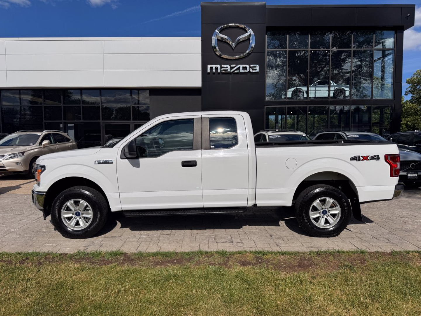 Ford F-150 pickup truck in white, extended cab, side view, parked in front of Mazda dealership building.