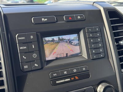 Ford F-150 center console with backup camera display showing parking grid and controls labeled RADIO, MEDIA, and keypad; climate/vehicle buttons around screen.