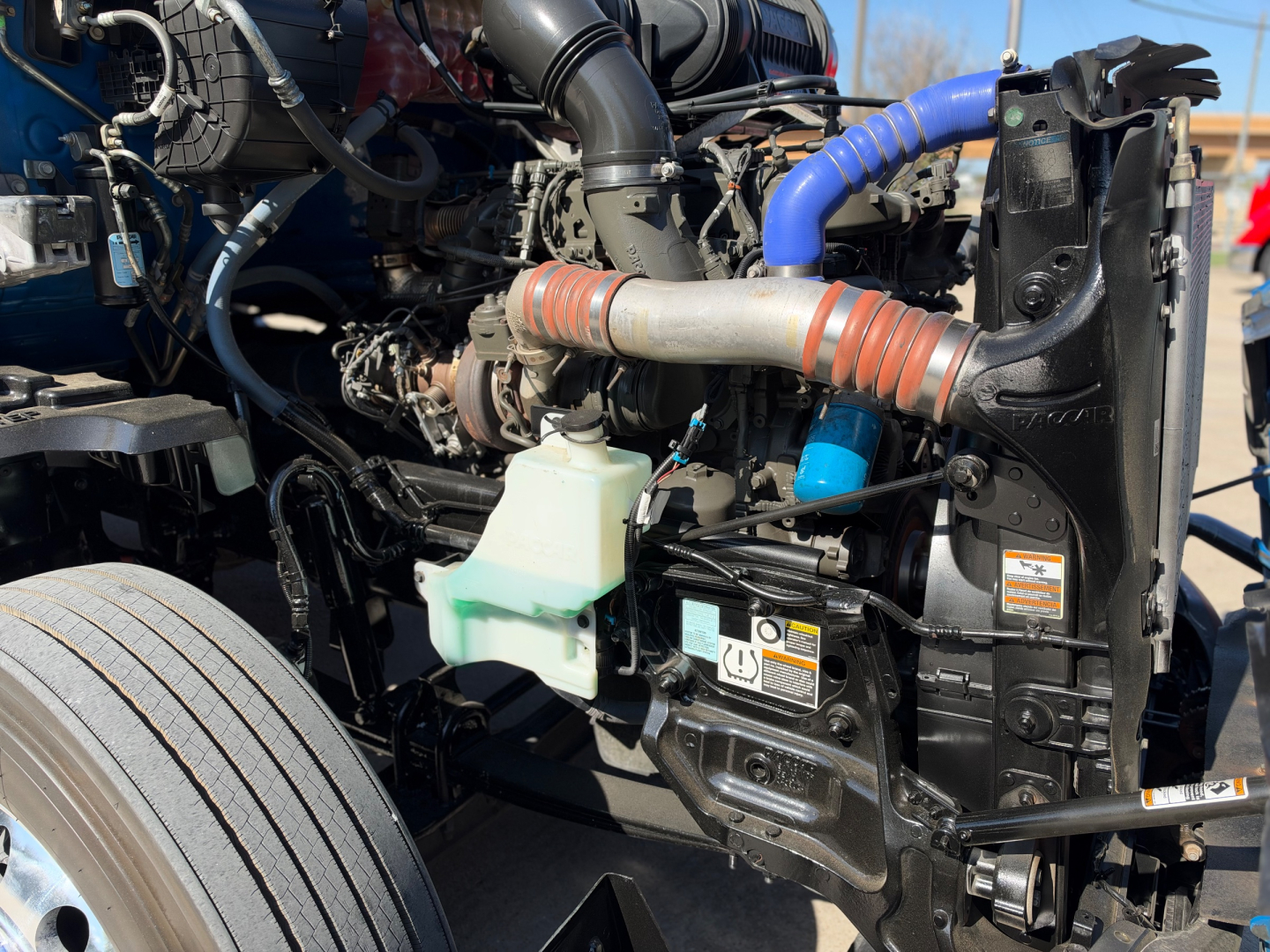 Kenworth T680 engine bay showing coolant reservoir, hoses, radiator support, belts, and wiring harness; visible labels and components in front of cab.
