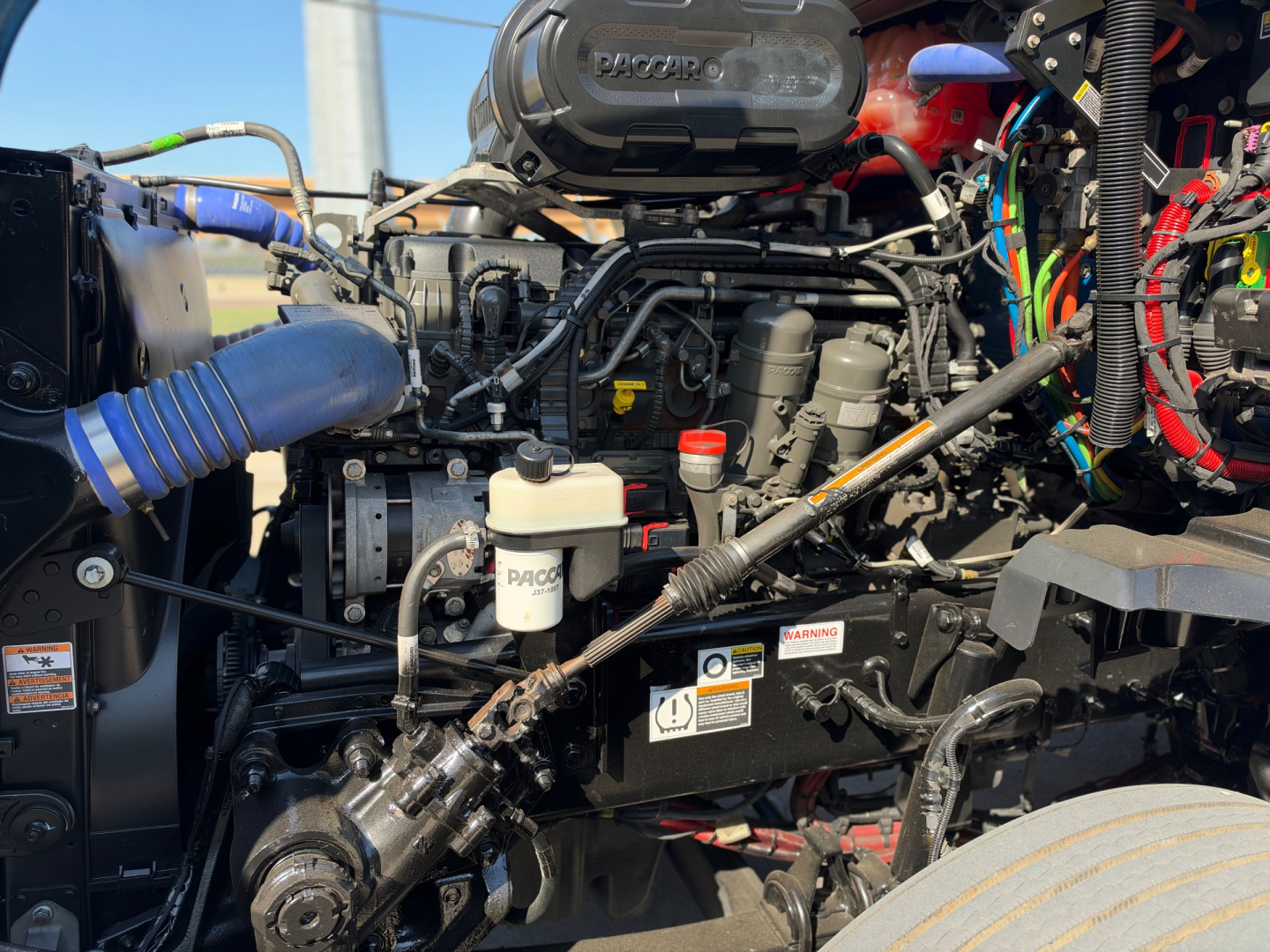 Kenworth T680 engine bay with visible components including air intake, hoses, wiring harness, reservoir caps, and label stickers.