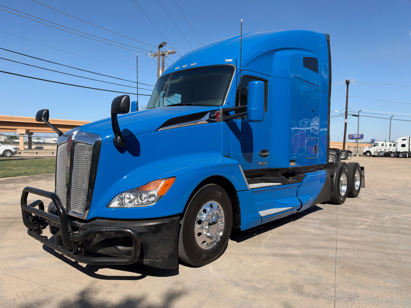 Blue Kenworth T680 semi-truck tractor in a dealership lot, front-left view, with bumper guard and chrome wheels.