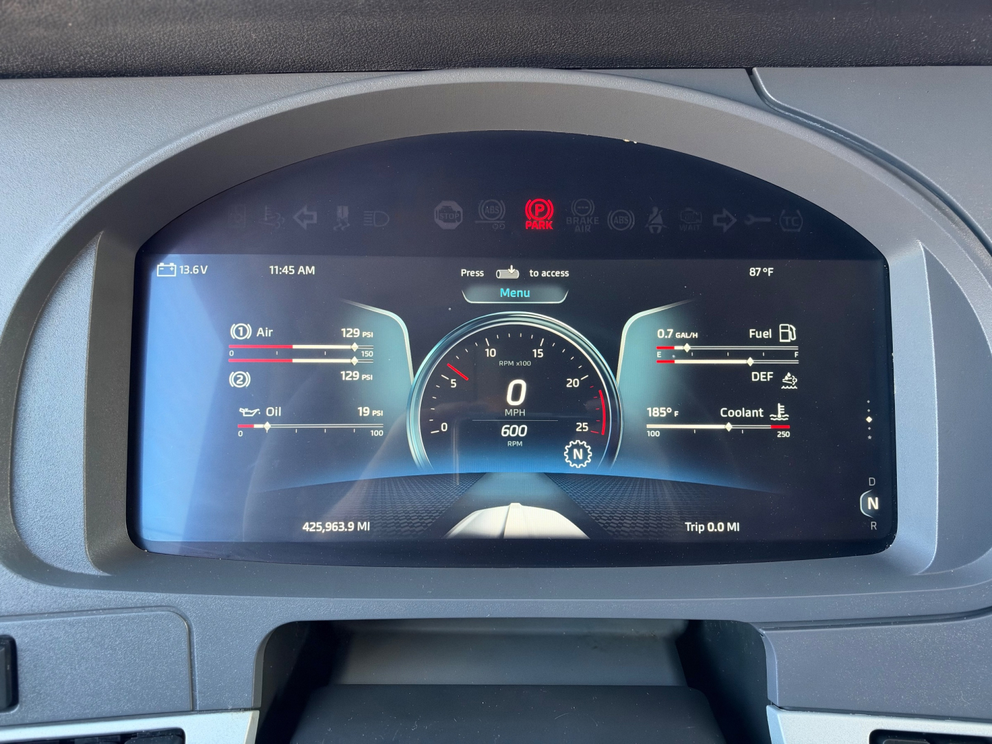 Kenworth T680 instrument cluster display showing around-dial digital gauges and trip data; center dial shows 0 mph with 600 RPM; left/right panels display fuel, DEF, oil, air and battery readouts.