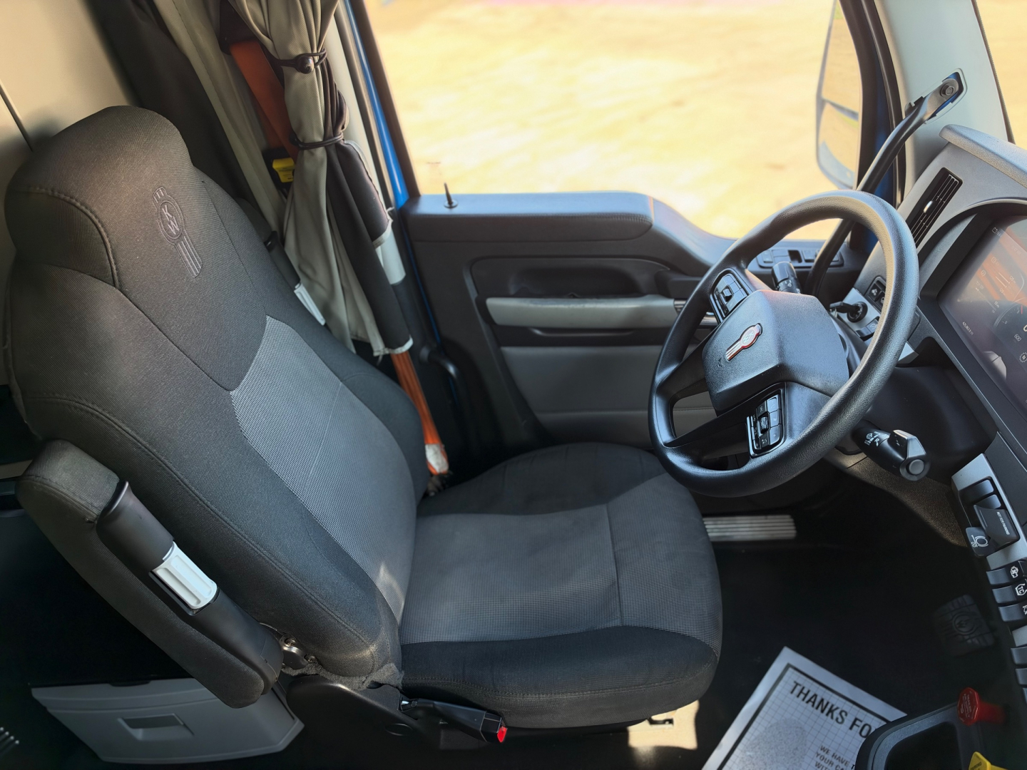 Kenworth T680 interior view showing driver seat, steering wheel, and dashboard from the cab with fabric seat and controls visible.