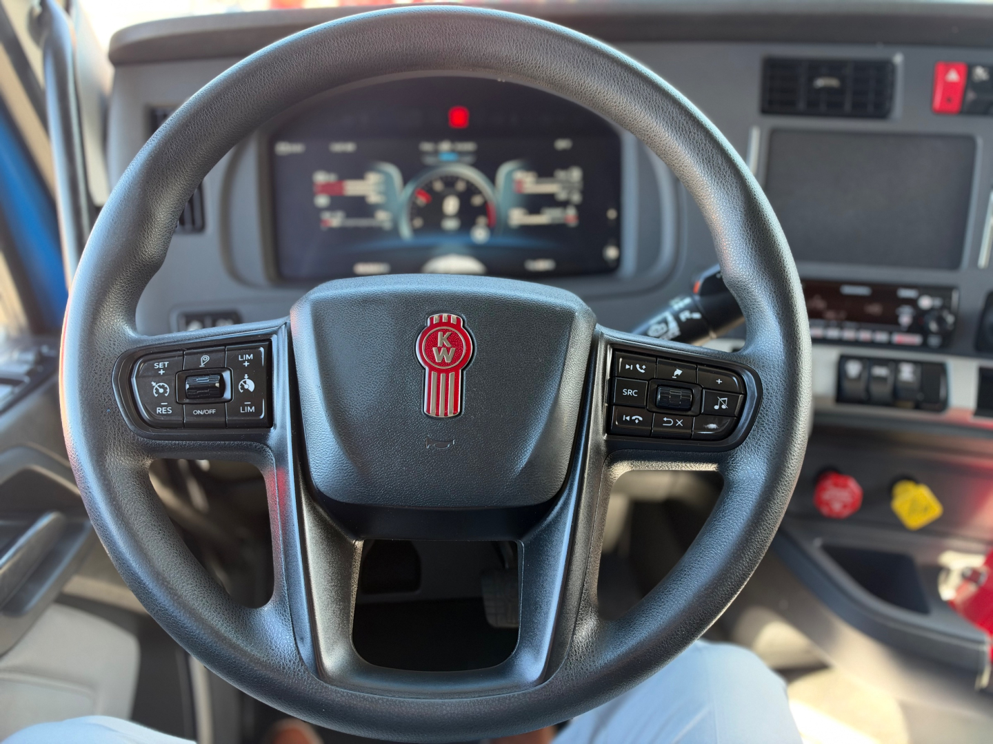 Kenworth T680 interior steering wheel and dashboard with digital gauges visible through steering wheel, Kenworth emblem at center, multiple control buttons on wheel, driver seat view.