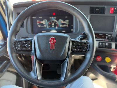 Kenworth T680 interior steering wheel and dashboard with digital gauges visible through steering wheel, Kenworth emblem at center, multiple control buttons on wheel, driver seat view.