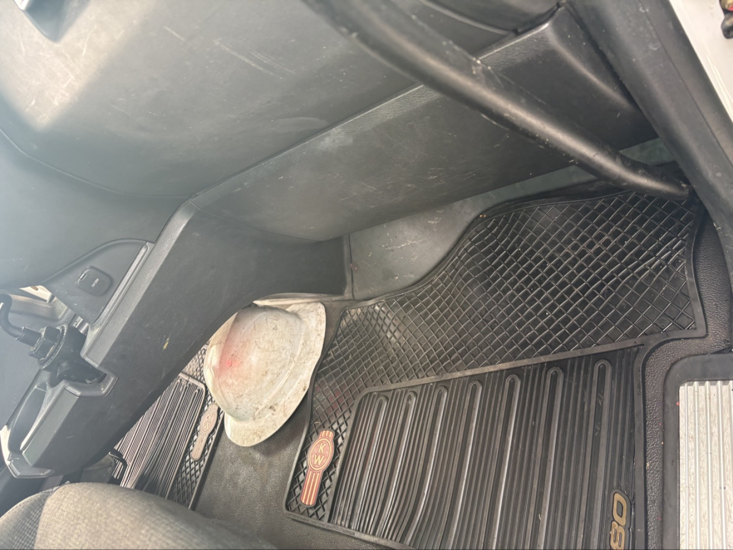 Interior dash area of a Kenworth T880 showing a floor mat and a deformed plastic kick panel near the passenger side; visible components include a red-tinted circular object and a portion of the dashboard.