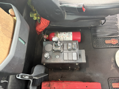 Kenworth T880 cab interior showing chassis area with a red fire extinguisher, a fuse/relay block, and pedals; visible floor mats and a badge with 'KW' emblem.