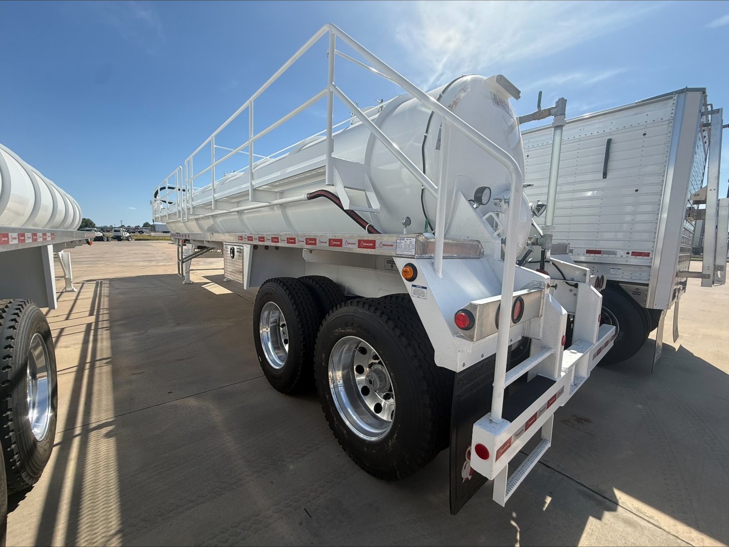 Trailer showing cylindrical tank with railings on top, tandem axle wheels, rear stairs and mud flaps. Labels and safety rails visible along frame.