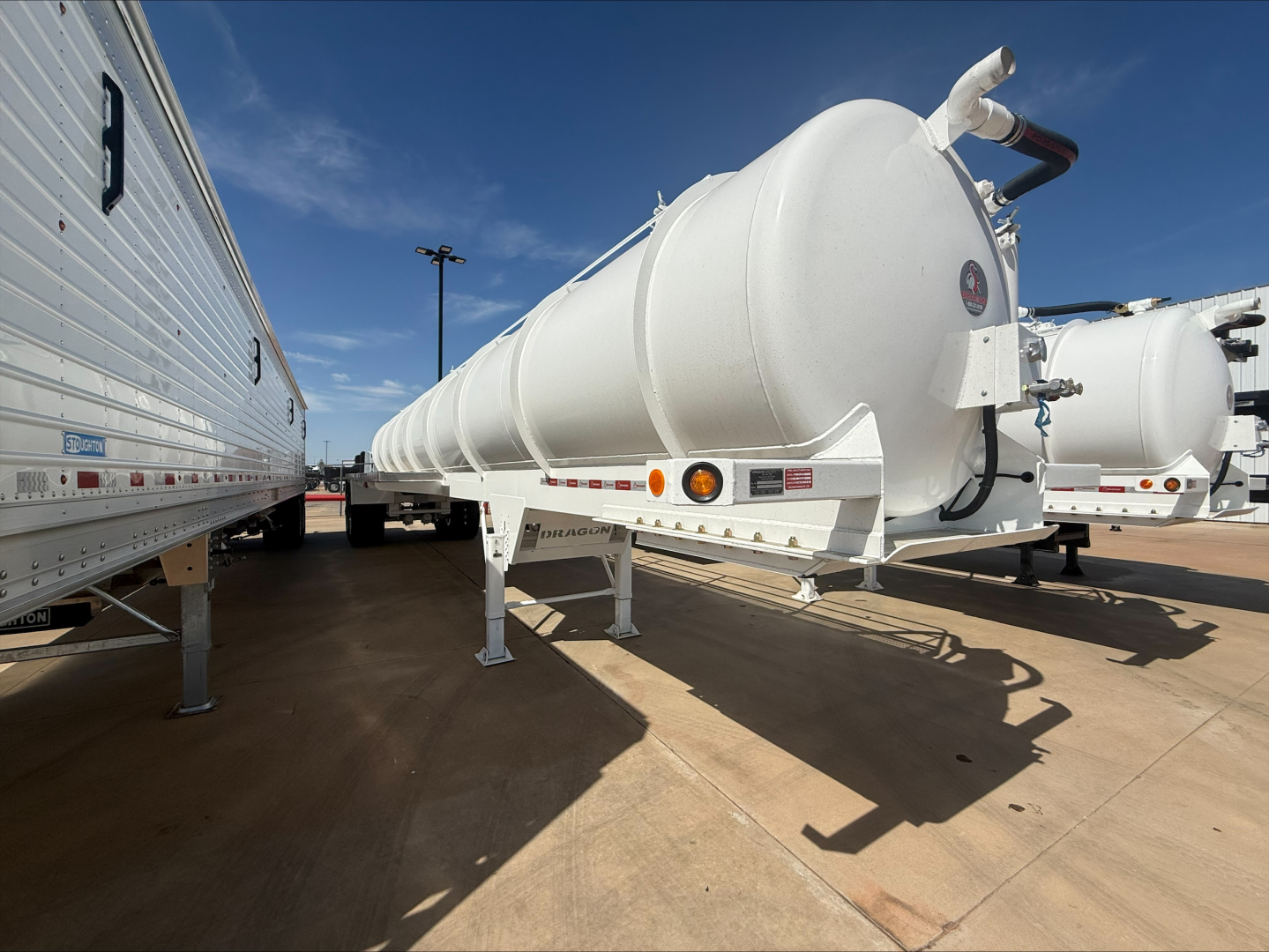 Vacuum trailer on a lot with long white cylindrical tank and ladder, showing support legs and tandem axles; visible text includes logos and warning labels on the side and rear.