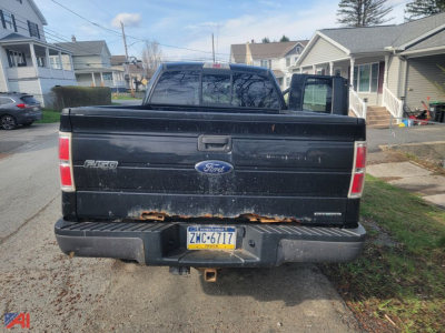 Ford F-150 tailgate and rear bumper on a black extended cab pickup, visible rust along the lower edge of the tailgate and bumper, license plate ZMC-6717, rural residential street background.