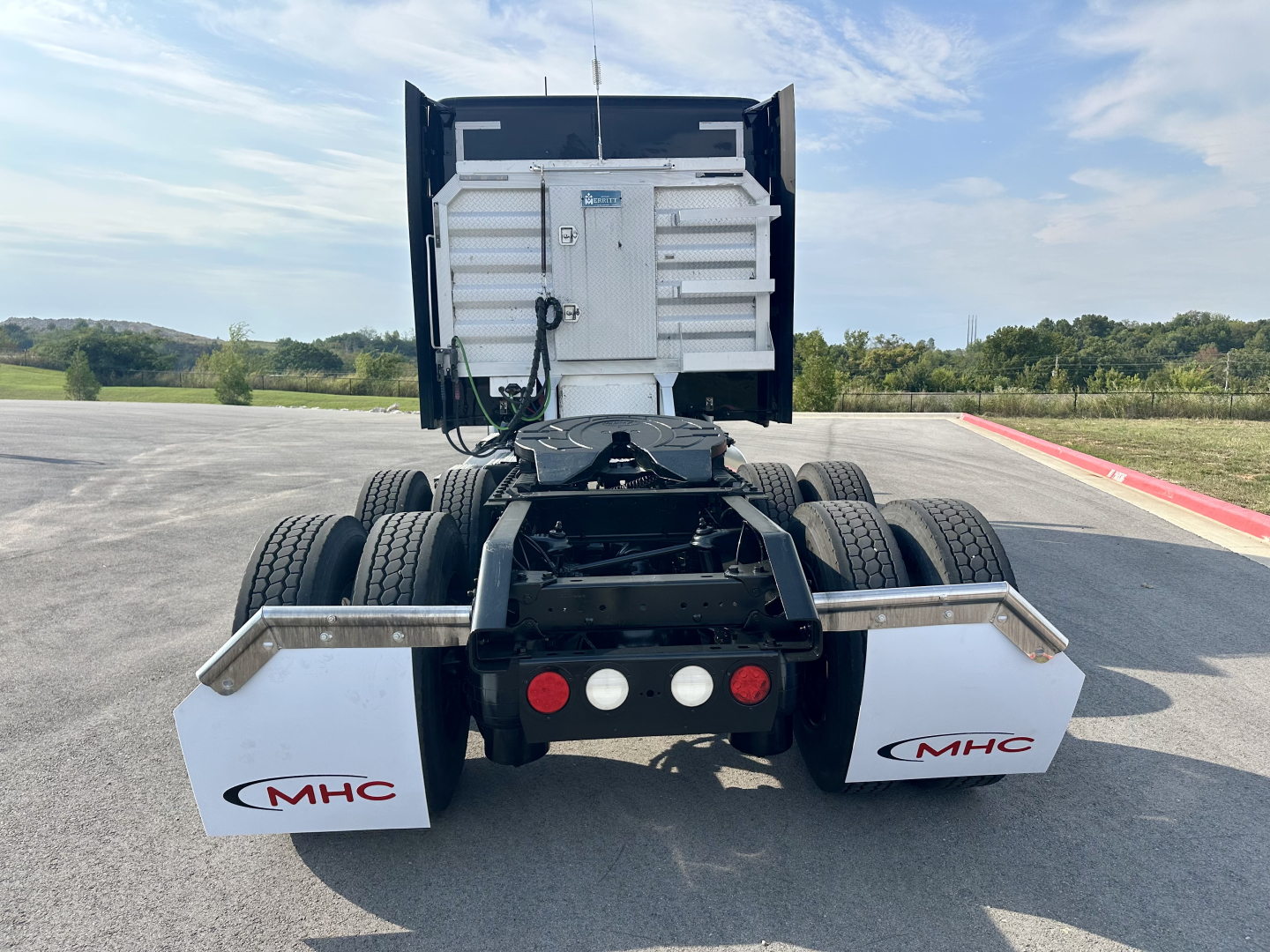 Kenworth T680 rear view showing sixth-wheel assembly and MHC white mud flaps with logos, truck chassis, and rear lights.