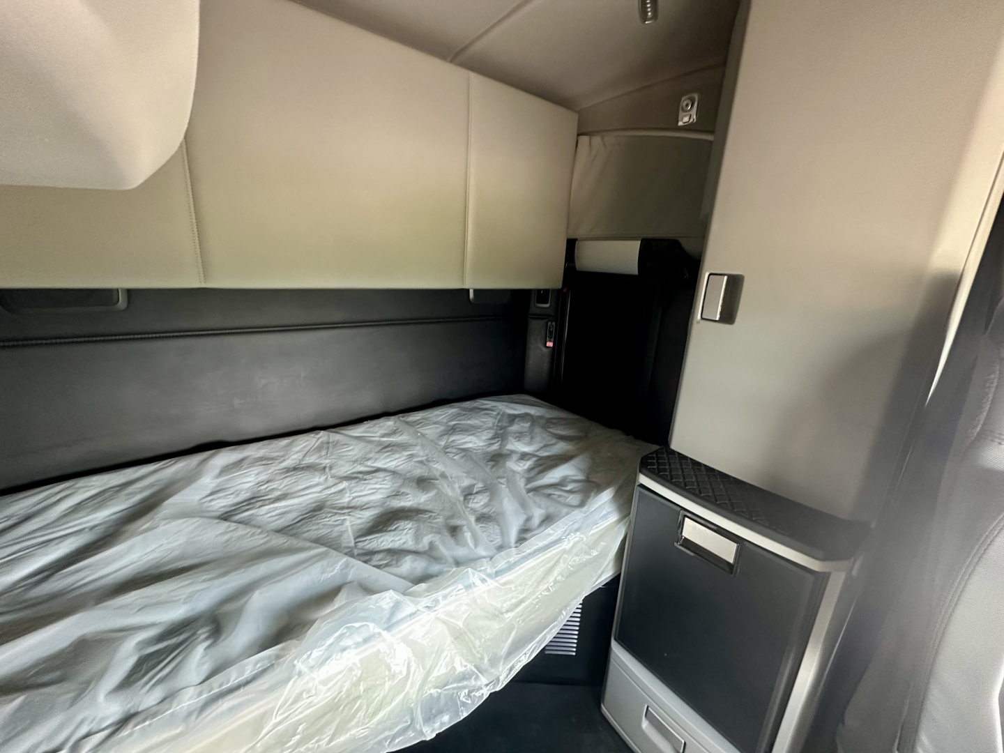 Truck sleeper cab interior with a wrapped mattress and light gray upholstery, storage cabinet, and side panel.
