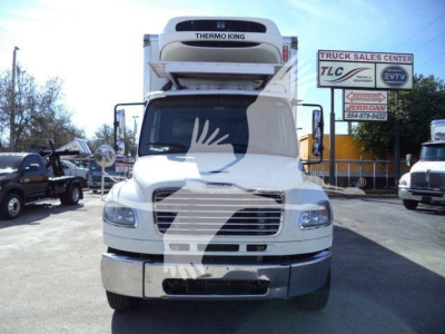 Front view of a white Freightliner M2 106 refrigerated truck with Thermo King unit on top. Text on signboards in background: 'TRUCK SALES CENTER' and 'TV' logos. No other legible labels on vehicle.