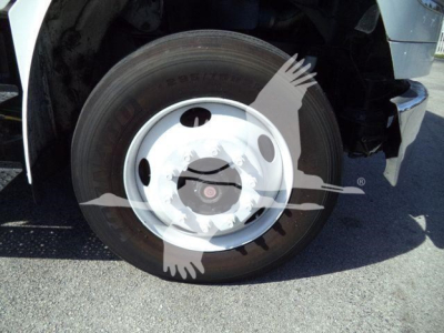 Wheel and tire of a Freightliner M2 106 front left with hub and rim visible, tire sidewall with faint lettering; part of the front fender and bumper area.