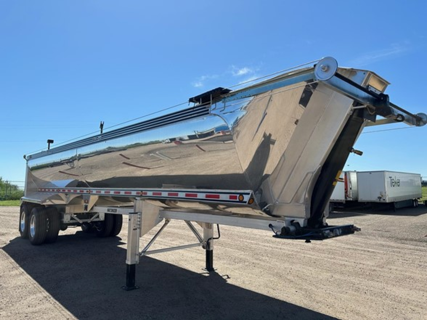Dump trailer with polished aluminum half-round end dump showing hydraulic cylinder and support legs, side railing, and tandem axles.
