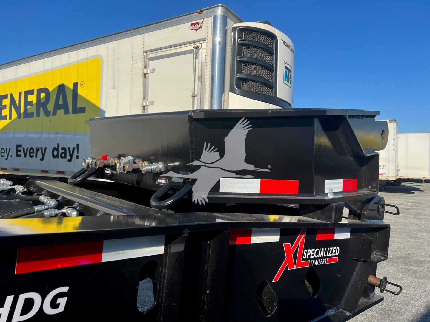 General view of a 2024 XL Specialized flip neck classic narrow trailer with a refrigeration unit on the front, black chassis with red-white safety markings, General trailer visible in background, MD36FB-488 stock.