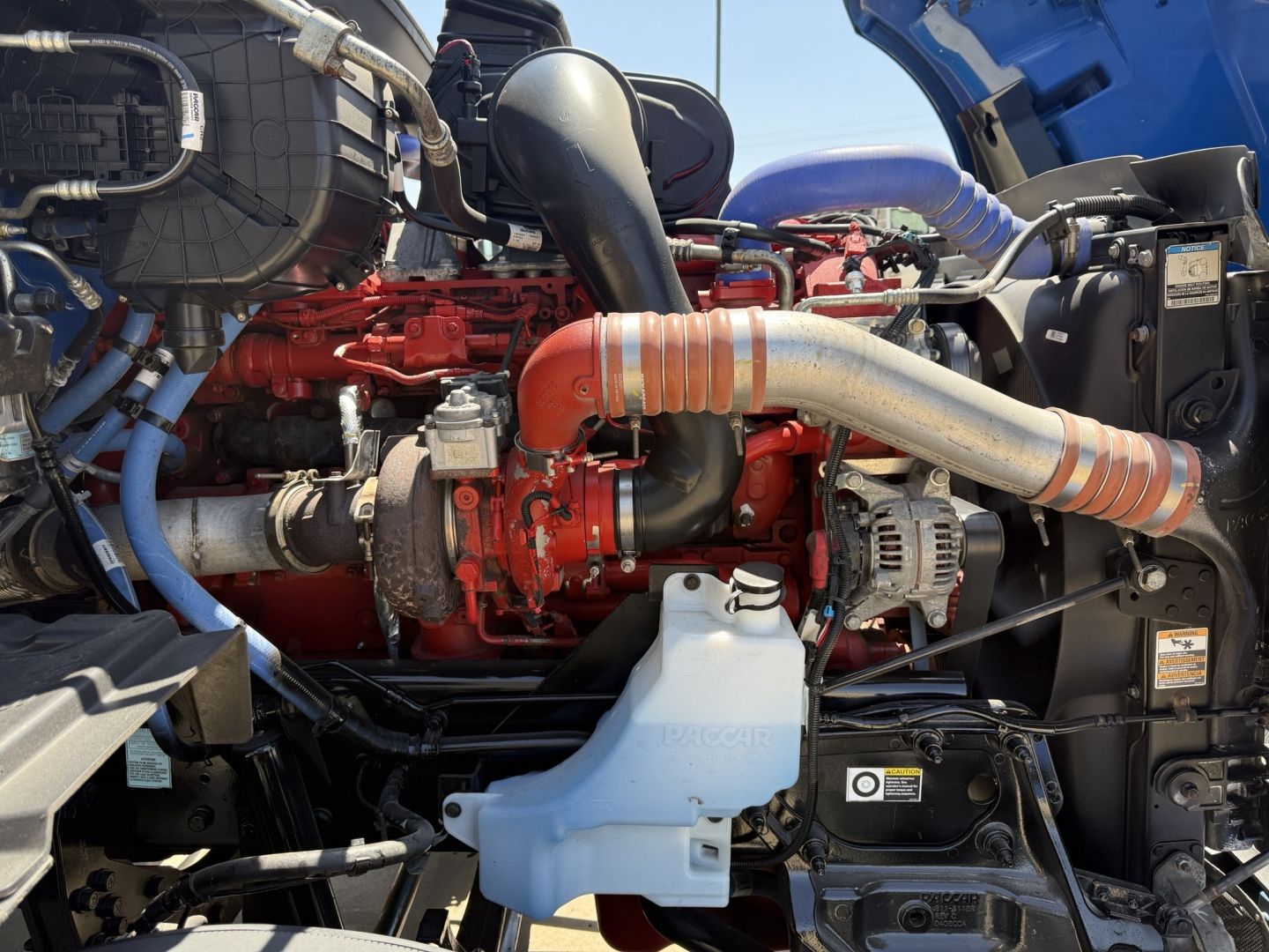 Peterbilt 579 engine bay showing red inline engine with large silver exhaust pipe, blue hoses, coolant reservoir, and various black plastic components.