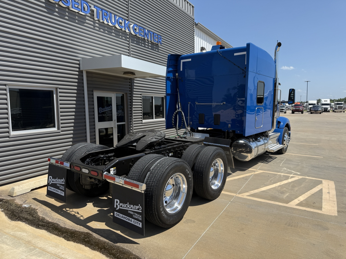 Peterbilt 579 tractor cab seen from rear side showing wheels and fifth wheel, blue cab, stock number B3387P and dealership building in background.