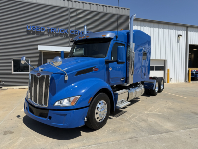 Peterbilt 579 sleeper cab truck in blue parked in front of dealership building, front three-quarter view showing chrome grille, dual fuel tanks, and polished wheels.