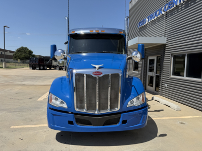Peterbilt 579 truck front view in blue with chrome grille and mirrors, highway skyline reflection, dealership building in background.