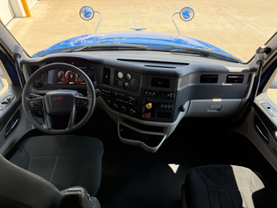 Interior of a Peterbilt truck cabin showing steering wheel, dashboard with gauges, center console, and driver seat; blue exterior hood visible from windshield.