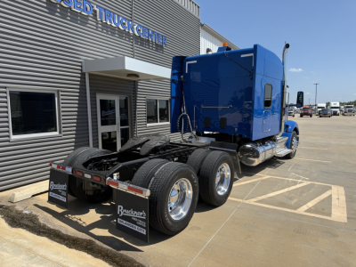 Peterbilt 579 tractor cab seen from rear side showing wheels and fifth wheel, blue cab, stock number B3387P and dealership building in background.