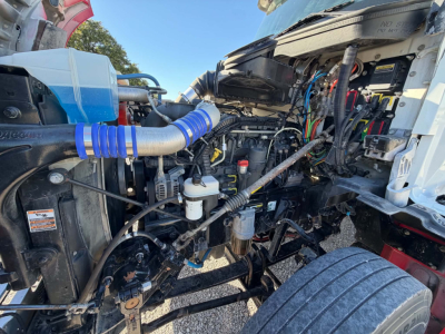Kenworth T880 engine bay showing assorted hoses, wiring, and components in view; visible coolant reservoir, air intake hose, and front wheel.