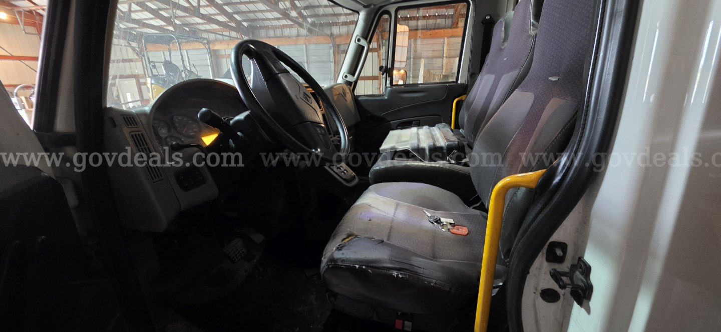 Truck cab interior showing steering wheel, driver and passenger seats, dash, and door. Visible wear on seat cushions and a yellow handrail near the seat.