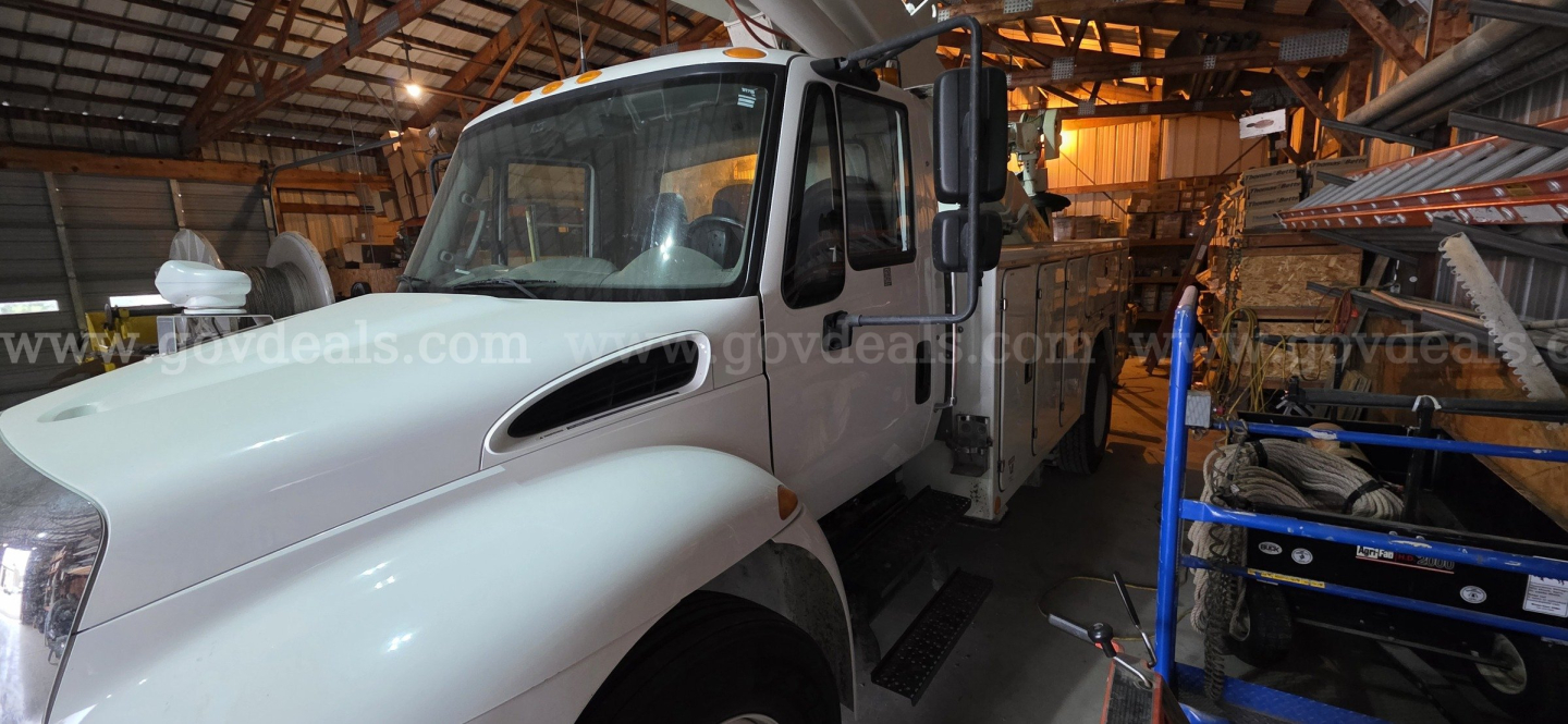 International 4300 boom truck in a workshop with white cab and equipment rack nearby.