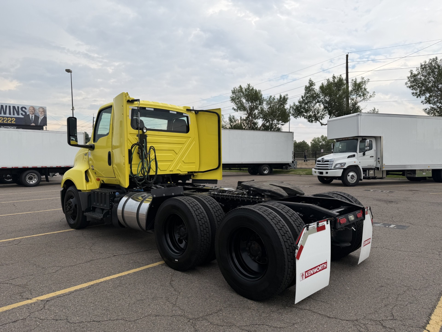 Truck chassis cab in a parking lot with long sleeper area missing; bright yellow International LT625 front cab, dual rear axles, white mud flaps, fuel tanks, and surrounding trucks.