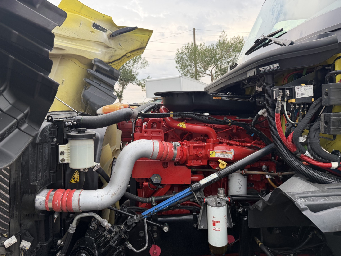 Truck engine bay of a 2023 International LT625 with red engine, white coolant/air hoses, and various black electrical harnesses; visible components include a white filter canister and red fan/engine accents.