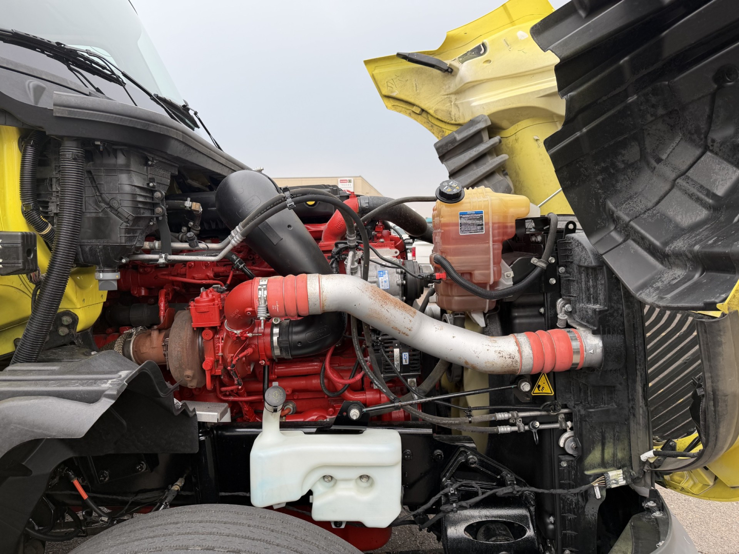 Engine bay of a 2023 International LT625 daytime view showing red engine, white coolant reservoir, and exposed hoses and wiring.