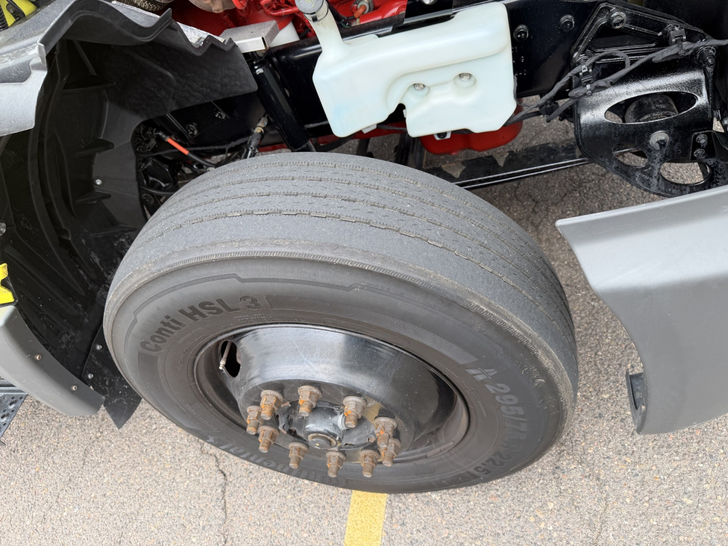 Wheel and front axle area of a used 2023 International LT625 conventional daycab showing a single front tire and hub with lug nuts; partial engine bay components and coolant reservoir visible.